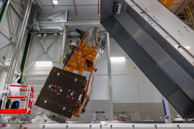 A satellite wrapped in golden thermal blankets and silver foil is suspended by cables in a clean room facility, with technicians in white protective suits visible below working on spacecraft assembly and testing.
