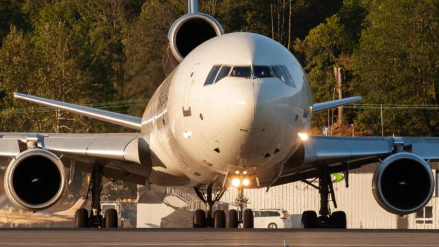 A white commercial aircraft sits on an airport tarmac with its landing lights illuminated, positioned directly facing the camera with trees and airport buildings visible in the background during golden hour lighting.