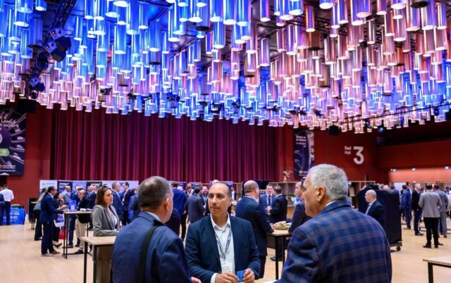Business professionals network and converse at a large conference or trade show featuring a striking ceiling installation of hundreds of illuminated cylindrical light fixtures in blue and purple hues. The modern venue has red curtained walls and displays, with attendees in formal business attire gathered around high-top tables throughout the spacious hall.