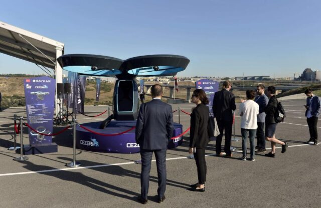 A blue aircraft or flying vehicle prototype on display at an outdoor exhibition, surrounded by formally dressed visitors and informational banners, with two large rotors visible on top of the futuristic-looking craft.
