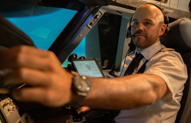 A pilot wearing a headset and white uniform shirt sits in an aircraft cockpit, operating flight controls with both hands while a tablet displaying flight information is visible nearby.