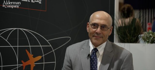 A bald man in glasses and a gray business suit with blue tie sits smiling in front of a dark wall featuring the Alderman & Company logo and a graphic of a globe with an airplane, suggesting an aviation or travel industry setting.