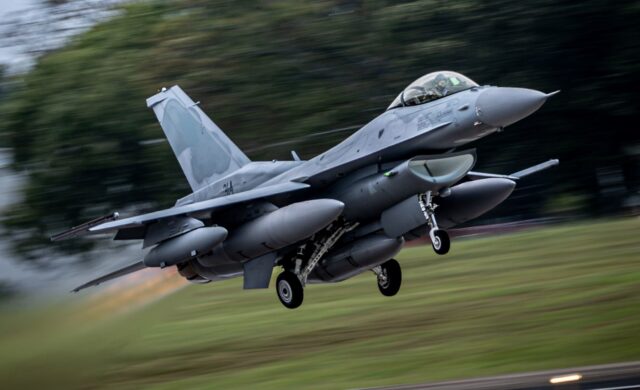 A military fighter jet takes off from a runway, with its landing gear extended and external fuel tanks or weapons pods visible under its wings. The aircraft appears to be an F-16 or similar single-engine fighter captured in motion against a blurred background of trees and grass.