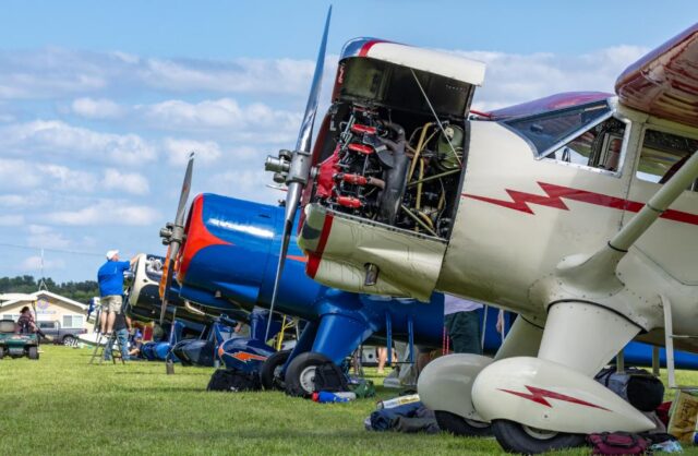 A vintage aircraft with its engine cowling removed sits on grass at what appears to be an airshow or aviation event, with maintenance personnel working on the exposed engine while other classic planes and spectators are visible in the background.