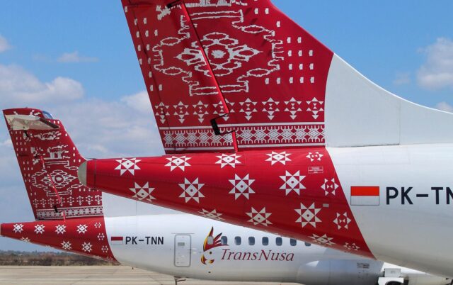 Two TransNusa aircraft tails featuring traditional Indonesian red and white geometric patterns, with registration codes PK-TNN and PK-TM visible on the white fuselages, parked on an airport tarmac under a blue sky with clouds.