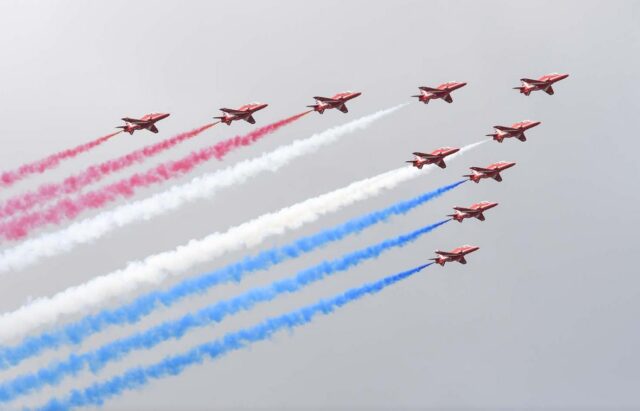 Nine red military aircraft fly in formation against a gray sky, releasing colored smoke trails in red, white, and blue that create parallel streams behind them during an aerobatic display.