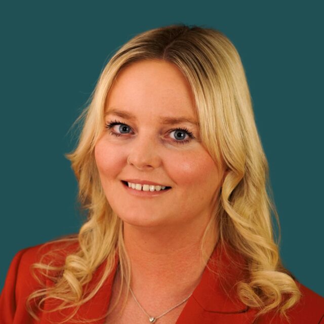 A professional headshot of a blonde woman wearing a red blazer and a delicate necklace, smiling warmly against a teal background.