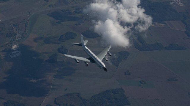 A commercial passenger aircraft flies high above a patchwork of agricultural fields and rural landscape, with white clouds visible in the distance below.