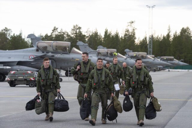 Military pilots in green flight suits walk across an airfield carrying equipment bags, with fighter jets visible in the background at what appears to be an air base.