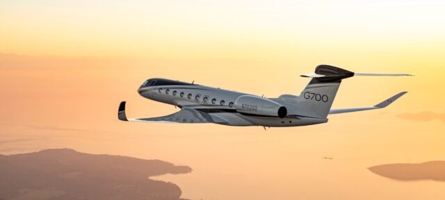 A Gulfstream G700 business jet flies through golden sunset skies above a mountainous coastline with islands visible below.