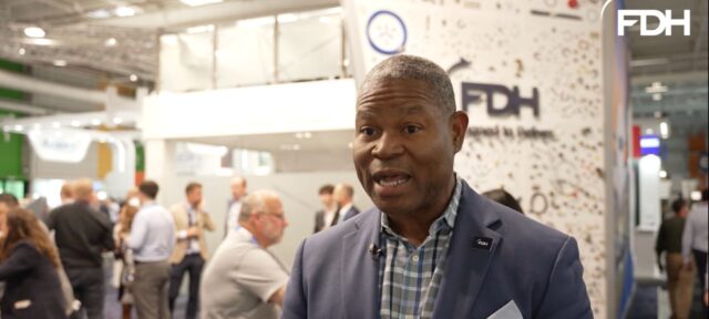 A professional man in a blue suit jacket and checkered shirt speaks at what appears to be a trade show or conference, with FDH signage and attendees visible in the background.