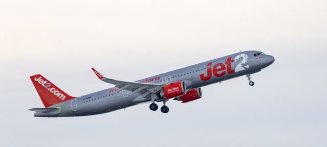 A Jet2.com aircraft in flight against a pale sky, featuring the airline's distinctive gray fuselage with red branding and tail design.