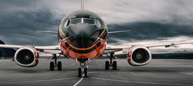 A commercial passenger aircraft with a distinctive black and orange livery sits on a runway under dramatic cloudy skies, viewed from the front showing its nose, engines, and landing gear.