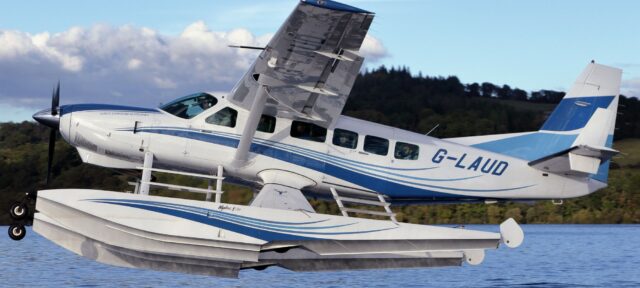 A white and blue seaplane with registration G-LAUD sits on calm water, featuring pontoon floats for water landing and takeoff capabilities against a backdrop of forested hills and cloudy sky.