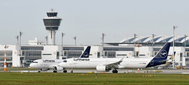 Two Lufthansa aircraft parked on the tarmac at an airport, with a modern control tower and terminal buildings visible in the background under an overcast sky.