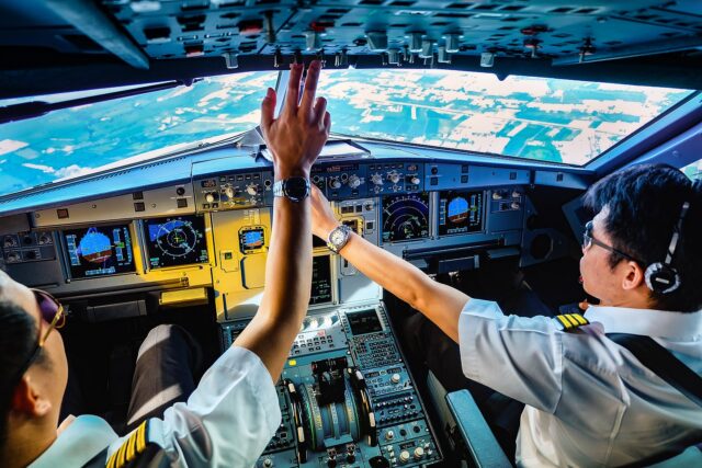 Two airline pilots in uniform celebrate with a high-five in the cockpit of a commercial aircraft, with complex instrument panels visible and an aerial view of land and water through the windscreen above them.