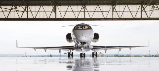 A sleek white private jet sits in a modern aircraft hangar with its reflection visible on the polished floor, positioned facing forward with landing gear deployed and engines mounted on the rear fuselage.