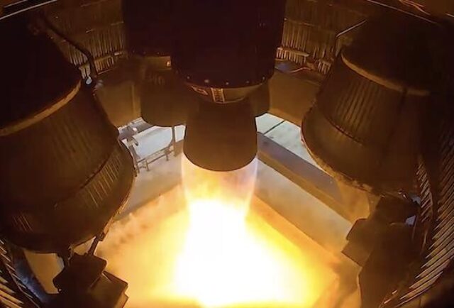 A rocket engine test firing produces bright orange flames and exhaust as viewed from above, showing the circular test facility structure surrounding the engine nozzle.