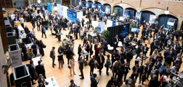 A crowded exhibition hall or trade show with hundreds of attendees in business attire networking around various vendor booths and displays in a large indoor venue with arched architecture.
