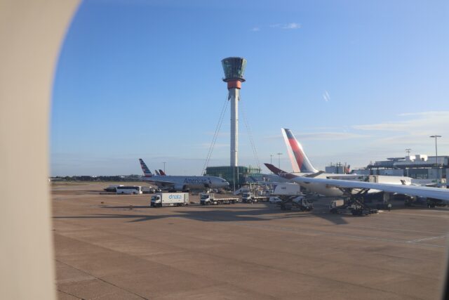 View of airport tarmac through aircraft window showing multiple commercial planes at gates, including American Airlines aircraft, with control tower and terminal buildings visible under clear blue sky.